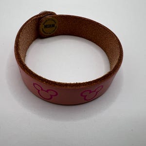 Puede incluir: Una pulsera de cuero rosa de tama&ntilde;o mediano con un borde marr&oacute;n. La pulsera presenta un patr&oacute;n repetido de siluetas rosas de Mickey Mouse. La pulsera se sujeta con una correa de cuero marr&oacute;n y un cierre met&aacute;lico.
