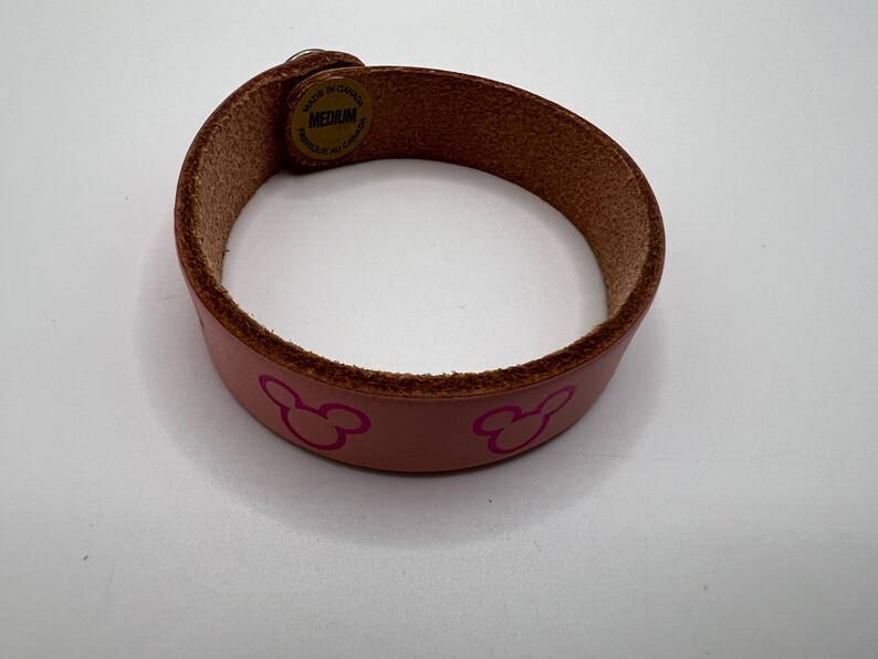Puede incluir: Una pulsera de cuero rosa de tama&ntilde;o mediano con un borde marr&oacute;n. La pulsera presenta un patr&oacute;n repetido de siluetas rosas de Mickey Mouse. La pulsera se sujeta con una correa de cuero marr&oacute;n y un cierre met&aacute;lico.