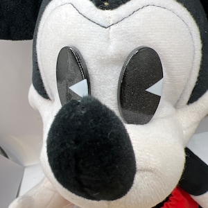 May include: A plush Mickey Mouse toy with a black nose, black and white face, and large black eyes with white accents. The toy has a red and yellow outfit and white gloves. The toy is a classic Disney character.