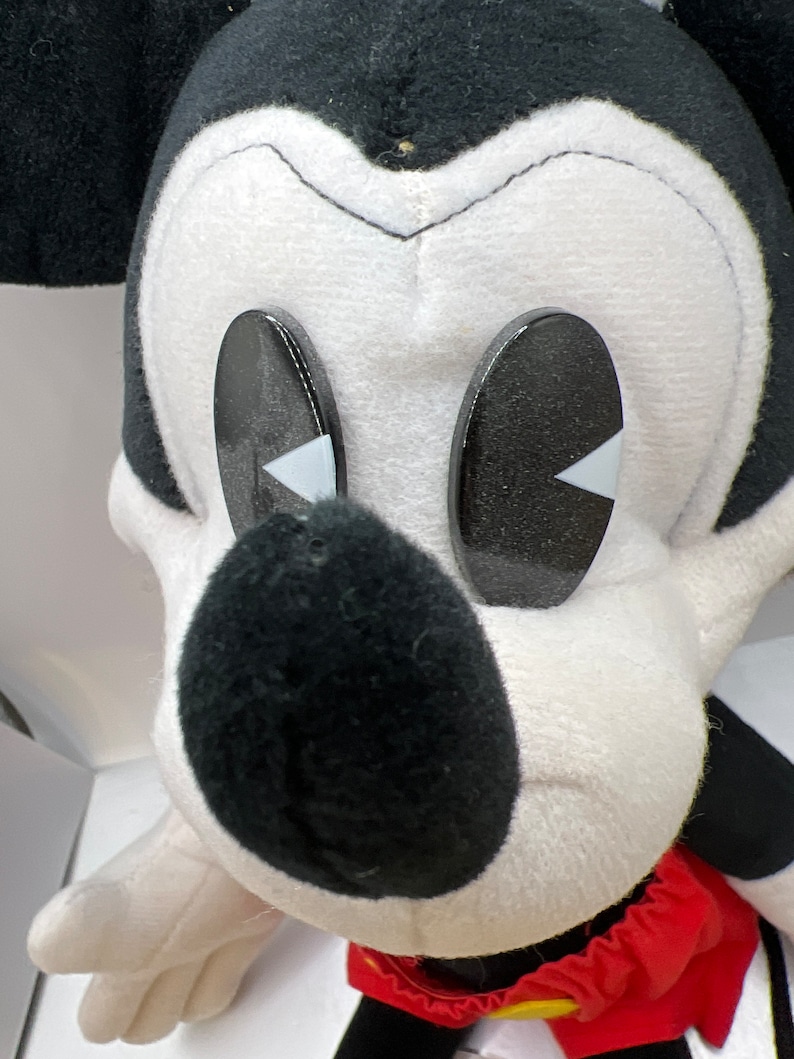May include: A plush Mickey Mouse toy with a black nose, black and white face, and large black eyes with white accents. The toy has a red and yellow outfit and white gloves. The toy is a classic Disney character.