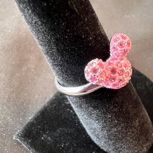 May include: A silver ring with a pink, crystal-covered Mickey Mouse design. The ring has a polished band, and the Mickey Mouse figure is made up of numerous small crystals, giving it a sparkly appearance. The ring is presented on a black velvet surface.