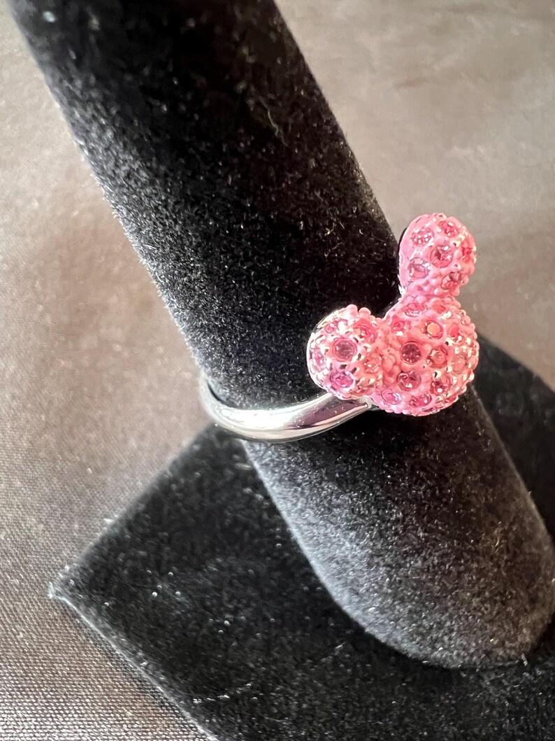 May include: A silver ring with a pink, crystal-covered Mickey Mouse design. The ring has a polished band, and the Mickey Mouse figure is made up of numerous small crystals, giving it a sparkly appearance. The ring is presented on a black velvet surface.