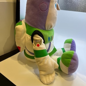 May include: A plush toy of Buzz Lightyear from the Toy Story franchise. The toy is sitting with its legs crossed and has a purple helmet, white and green spacesuit, and a yellow and black "Laser" patch on its arm.