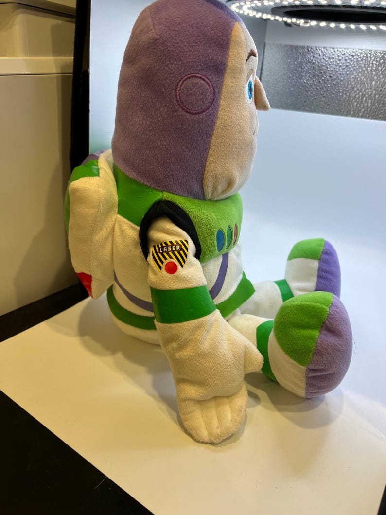 May include: A plush toy of Buzz Lightyear from the Toy Story franchise. The toy is sitting with its legs crossed and has a purple helmet, white and green spacesuit, and a yellow and black "Laser" patch on its arm.
