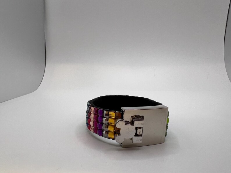 Puede incluir: Una pulsera ancha de cuero negro con filas de cuentas de colores rosa, morado, amarillo y verde. La pulsera tiene un cierre plateado con un dise&ntilde;o de Mickey Mouse.