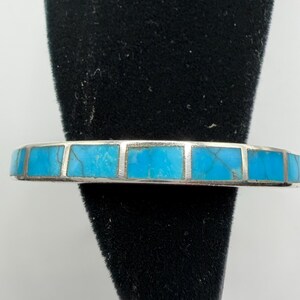 May include: A silver bracelet featuring a row of rectangular, bright blue turquoise inlays with dark veining. The metal band is polished and smooth, holding the individual stone segments in a clean, linear arrangement across the front of the piece.