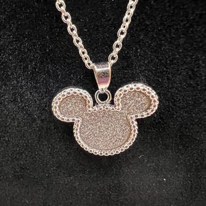 K&ouml;nnte beinhalten: Silberfarbene Halskette mit einem Anh&auml;nger in Form eines Mickey Mouse-Kopfes. Der Anh&auml;nger hat eine strukturierte Oberfl&auml;che und einen Perlenrand. Die Kette ist im filigranen Glieder-Stil gehalten. Der Hintergrund ist schwarz.