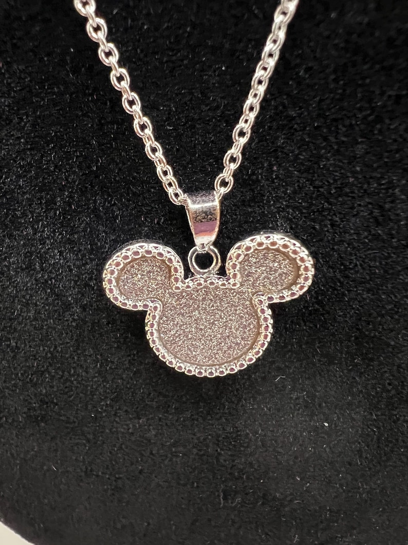 K&ouml;nnte beinhalten: Silberfarbene Halskette mit einem Anh&auml;nger in Form eines Mickey Mouse-Kopfes. Der Anh&auml;nger hat eine strukturierte Oberfl&auml;che und einen Perlenrand. Die Kette ist im filigranen Glieder-Stil gehalten. Der Hintergrund ist schwarz.