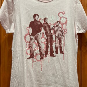 May include: A pink t-shirt with a graphic print of three men in a band. The band's name is "Rascal Flatts" and is printed in white on the shirt. The graphic is in shades of brown and pink.