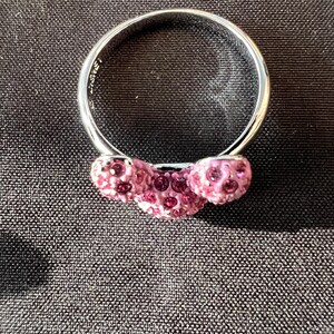 May include: A silver ring featuring three pink crystal spheres. The ring is set against a dark backdrop, accentuating the sparkle of the crystals. The band is smooth and polished.