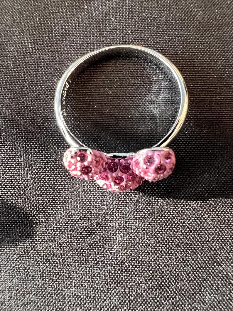May include: A silver ring featuring three pink crystal spheres. The ring is set against a dark backdrop, accentuating the sparkle of the crystals. The band is smooth and polished.