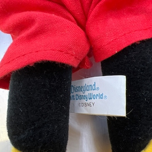 May include: Close-up of a plush toy's legs and red outfit. The legs are black with yellow feet. The red outfit is visible, and a tag reads "Disneyland, Walt Disney World, &copy;DISNEY."