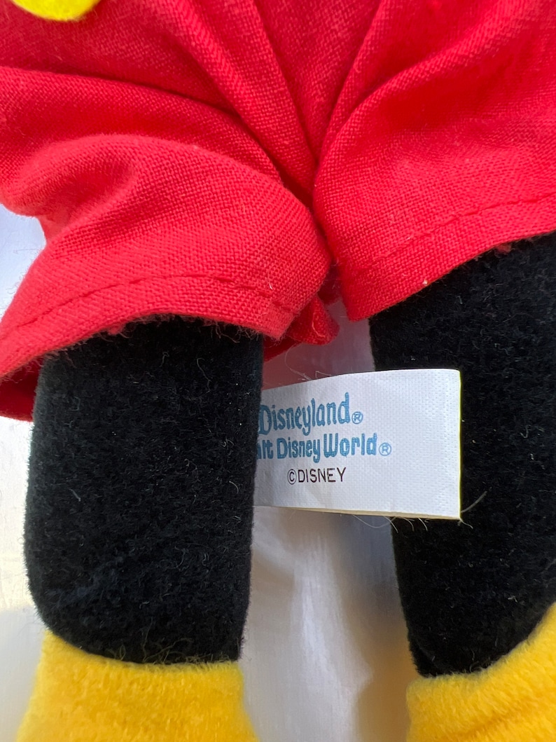 May include: Close-up of a plush toy's legs and red outfit. The legs are black with yellow feet. The red outfit is visible, and a tag reads "Disneyland, Walt Disney World, &copy;DISNEY."