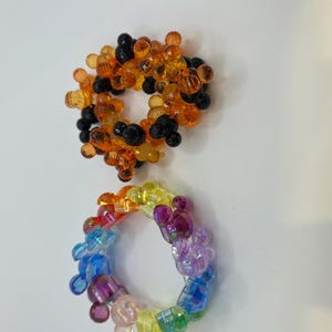 Puede incluir: Dos pulseras de cuentas. La pulsera superior presenta cuentas naranjas y negras. La pulsera inferior es un arco&iacute;ris de colores, incluyendo azul, morado, amarillo y verde. Ambas pulseras son circulares.