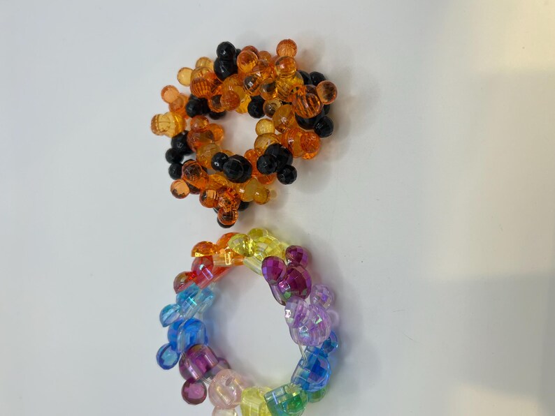 Puede incluir: Dos pulseras de cuentas. La pulsera superior presenta cuentas naranjas y negras. La pulsera inferior es un arco&iacute;ris de colores, incluyendo azul, morado, amarillo y verde. Ambas pulseras son circulares.