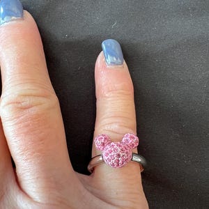 May include: A silver ring with a pink crystal Mickey Mouse head design. The ring's band is silver, and the Mickey Mouse head is made of small, sparkling pink crystals. The ring is worn on a finger. The ring is a piece of jewellery.