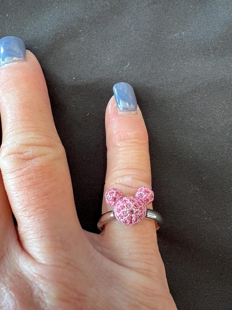 May include: A silver ring with a pink crystal Mickey Mouse head design. The ring's band is silver, and the Mickey Mouse head is made of small, sparkling pink crystals. The ring is worn on a finger. The ring is a piece of jewellery.