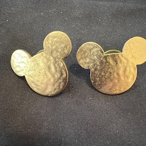 Pu&ograve; includere: Due orecchini dorati a forma di Mickey Mouse. Ogni orecchino ha una superficie strutturata e un perno per orecchie forate. Gli orecchini sono esposti su uno sfondo di tessuto nero.