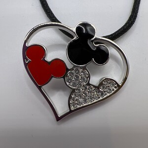 Puede incluir: Un collar con un colgante de coraz&oacute;n plateado con un dise&ntilde;o de Mickey Mouse en negro y rojo. El colgante est&aacute; adornado con brillantes transparentes.