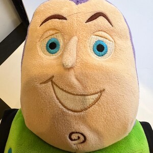 May include: A plush toy of Buzz Lightyear from the Toy Story franchise. The toy has a purple head with a smiling face, blue eyes, and a green and yellow body. The toy has a yellow name tag that reads "LIGHTYEAR".