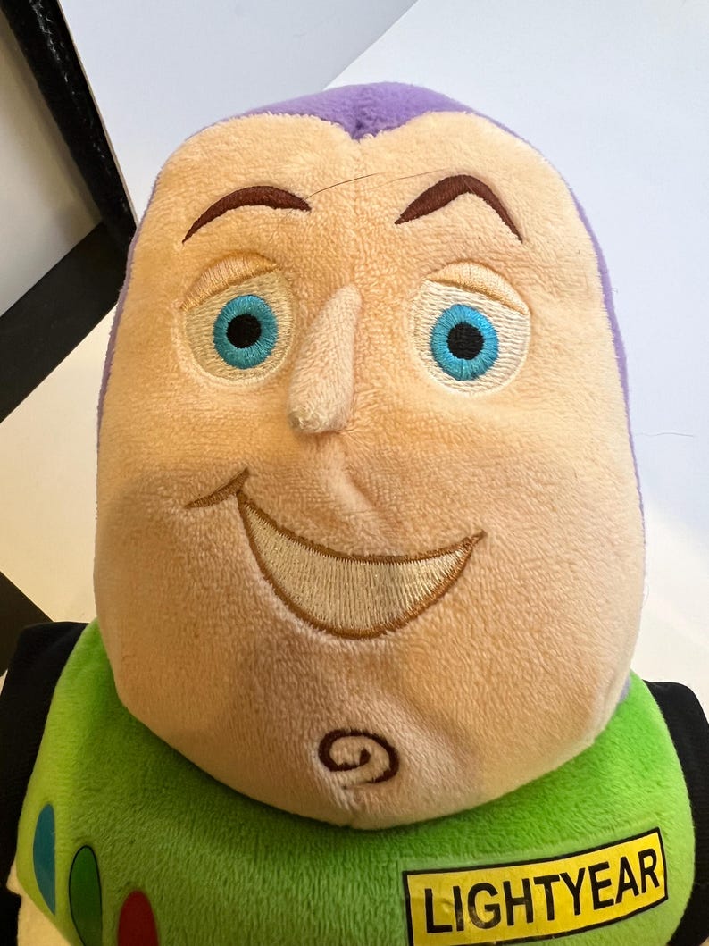 May include: A plush toy of Buzz Lightyear from the Toy Story franchise. The toy has a purple head with a smiling face, blue eyes, and a green and yellow body. The toy has a yellow name tag that reads "LIGHTYEAR".