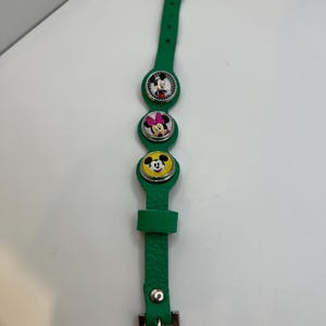 Puede incluir: Pulsera de cuero verde con tres colgantes circulares con Mickey Mouse, Minnie Mouse y el Pato Donald. La pulsera tiene un cierre de hebilla y est&aacute; dise&ntilde;ada para un ni&ntilde;o.