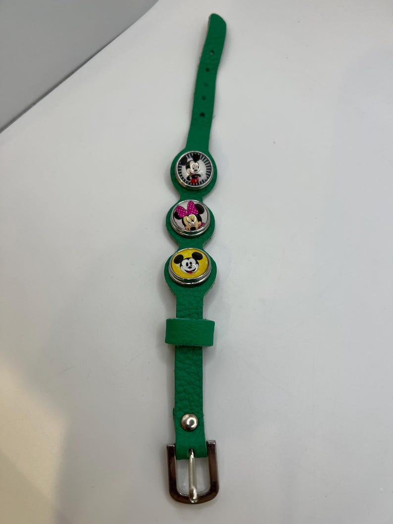 Puede incluir: Pulsera de cuero verde con tres colgantes circulares con Mickey Mouse, Minnie Mouse y el Pato Donald. La pulsera tiene un cierre de hebilla y est&aacute; dise&ntilde;ada para un ni&ntilde;o.