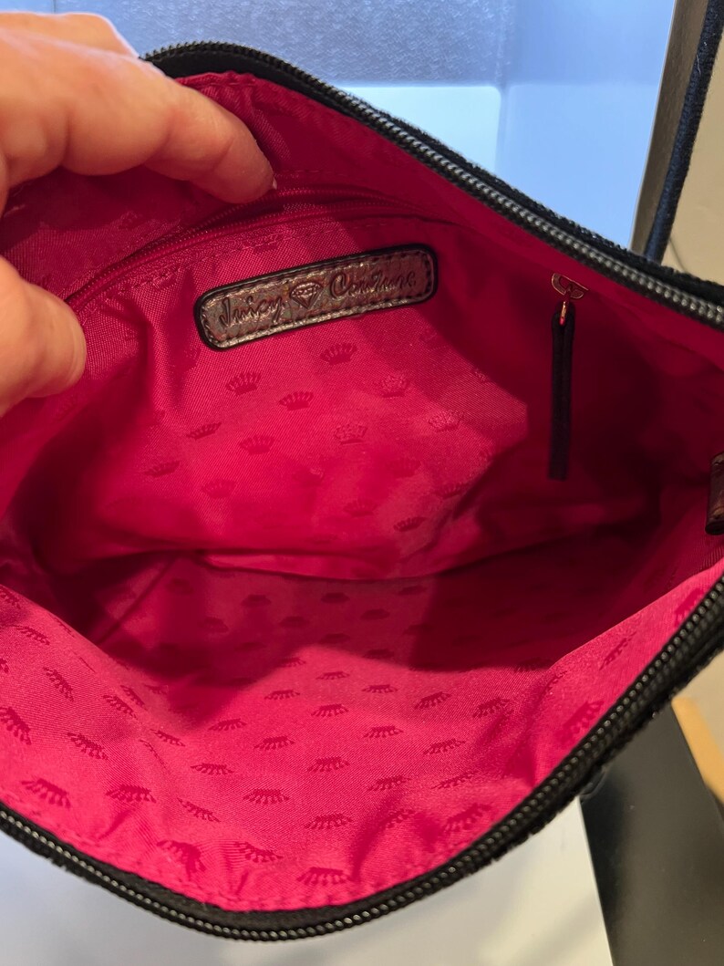 May include: The inside of a black purse with a pink lining. The lining has a repeating pattern of crowns. The purse has a silver tag that says "Juicy Couture".