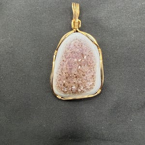 May include: A teardrop-shaped pendant featuring a central cluster of light purple druzy crystals surrounded by a smooth, white, banded agate border. The stone is encased in a gold-colored wire-wrapped frame with a matching wire bail at the top.