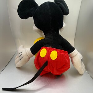 May include: A plush Mickey Mouse toy, viewed from the back. The toy features a black head and ears, white gloves, and red shorts with yellow buttons. A black tail extends from the shorts. The toy is set against a light background.