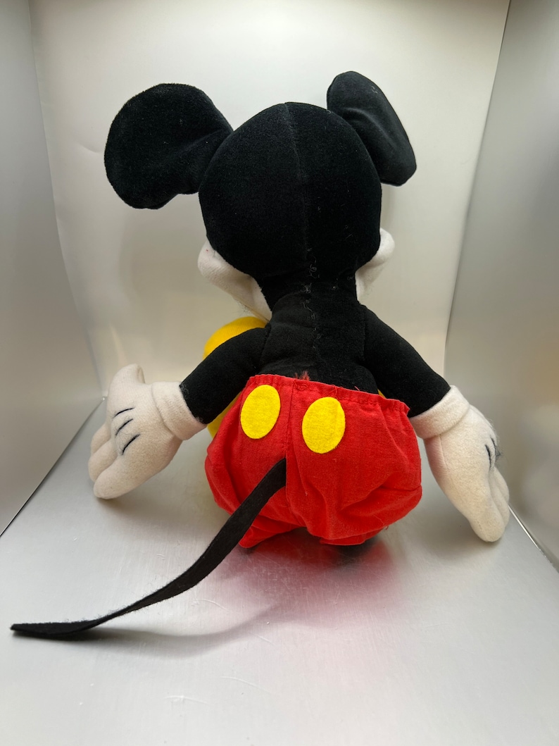 May include: A plush Mickey Mouse toy, viewed from the back. The toy features a black head and ears, white gloves, and red shorts with yellow buttons. A black tail extends from the shorts. The toy is set against a light background.