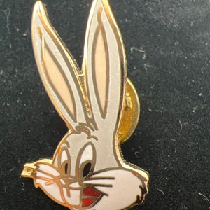May include: A gold-colored pin featuring Bugs Bunny's head. The pin is white with black eyes and a red mouth. The ears are long and white with a gold outline.