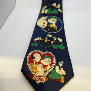 May include: Navy blue tie featuring Popeye the Sailor Man and Olive Oyl cartoon graphics. The tie has various scenes, including a heart-shaped image with the text "First Cola" and the word "MEN!" in a speech bubble.