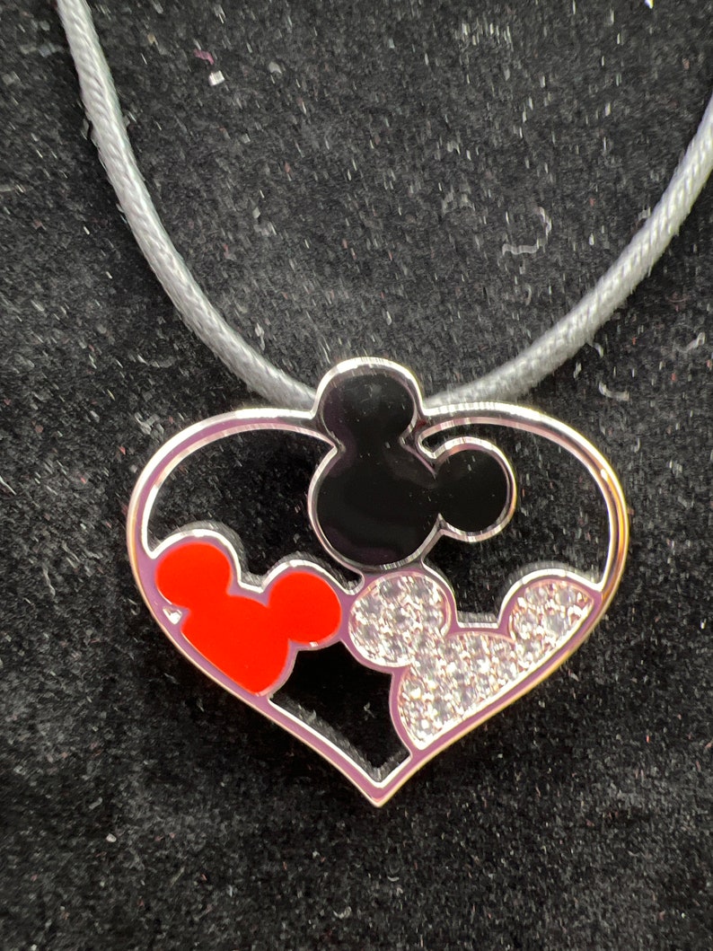 Puede incluir: Un collar con un colgante en forma de coraz&oacute;n plateado con tres cabezas de Mickey Mouse. Las cabezas de Mickey Mouse son negras, rojas y transparentes con diamantes de imitaci&oacute;n.