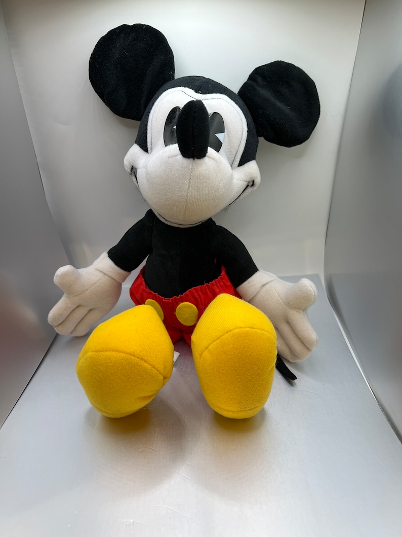 May include: A plush Mickey Mouse toy, featuring a black body, ears, and arms, with white gloves and face. The toy has red shorts with yellow buttons and large yellow feet. A classic Disney character.