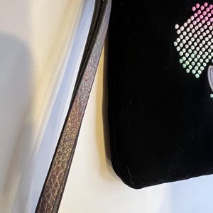May include: A black velvet clutch bag with a rainbow-colored rhinestone design. The bag has a long, adjustable strap.