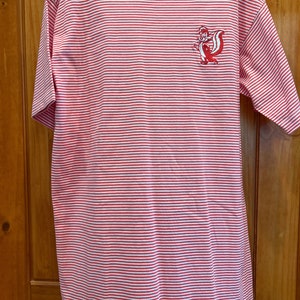 May include: Red and white striped t-shirt with a small embroidered cartoon skunk on the chest. The shirt has a crew neck and short sleeves. The shirt is hanging on a wooden door.