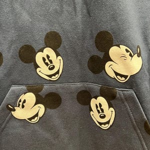 Pu&ograve; includere: Una felpa blu con un motivo ripetitivo di volti di Topolino. La felpa ha una tasca a marsupio.