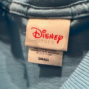 May include: A white tag with red text that reads "Disney Store" and "100% cotton/coton assembled in El Salvador of U.S.A. fabric" and "Small".