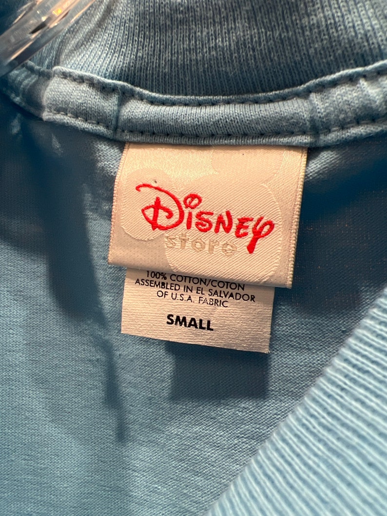 May include: A white tag with red text that reads "Disney Store" and "100% cotton/coton assembled in El Salvador of U.S.A. fabric" and "Small".