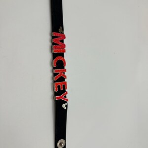 Puede incluir: Pulsera negra con el nombre "MICKEY" en letras rojas con purpurina. La pulsera tiene cierres de bot&oacute;n plateados y un bot&oacute;n plateado en la parte superior. Las letras est&aacute;n delineadas en plata.