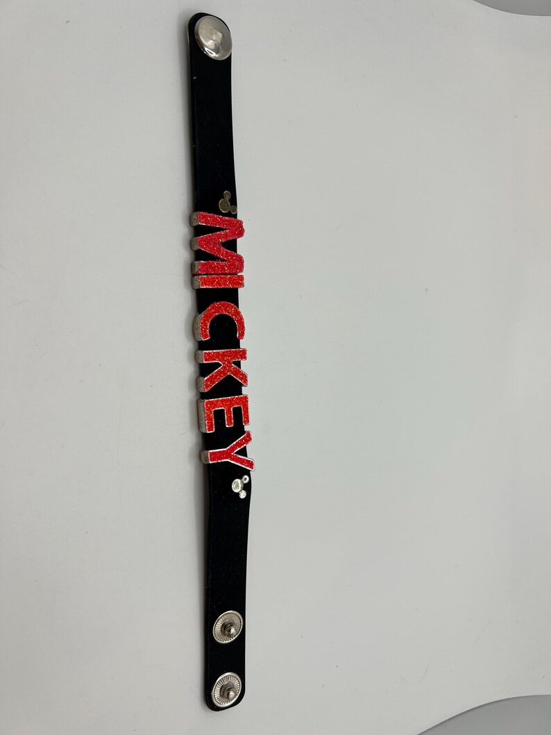 Puede incluir: Pulsera negra con el nombre "MICKEY" en letras rojas con purpurina. La pulsera tiene cierres de bot&oacute;n plateados y un bot&oacute;n plateado en la parte superior. Las letras est&aacute;n delineadas en plata.