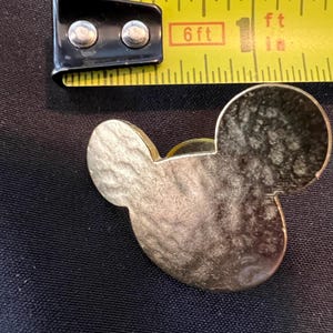 Pu&ograve; includere: Una spilla a forma di testa di Topolino color oro. La spilla &egrave; accanto a un metro a nastro giallo, che mostra 2,5 cm. La spilla ha una superficie strutturata e si trova su uno sfondo nero.