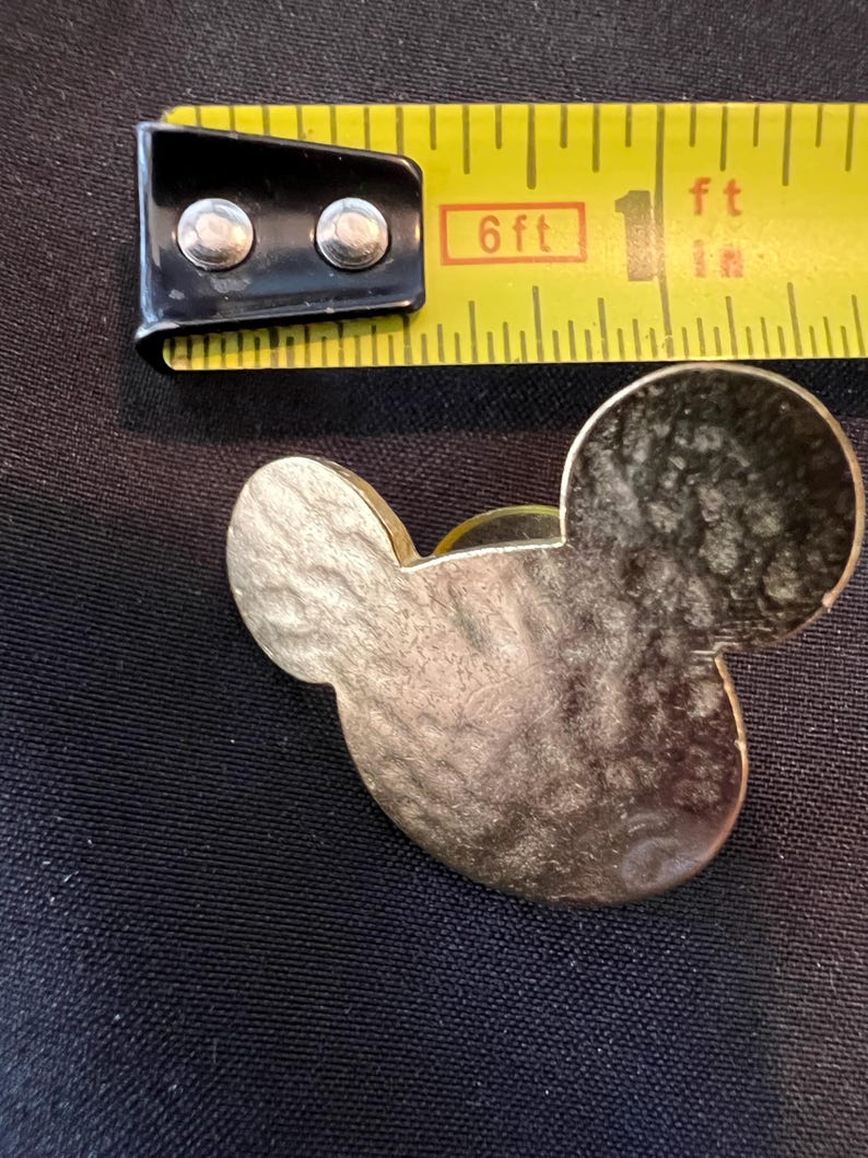 Pu&ograve; includere: Una spilla a forma di testa di Topolino color oro. La spilla &egrave; accanto a un metro a nastro giallo, che mostra 2,5 cm. La spilla ha una superficie strutturata e si trova su uno sfondo nero.