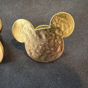 Pu&ograve; includere: Spilla dorata a forma di testa di Topolino con superficie strutturata. La spilla ha due orecchie circolari e una testa arrotondata. La spilla &egrave; su uno sfondo scuro.