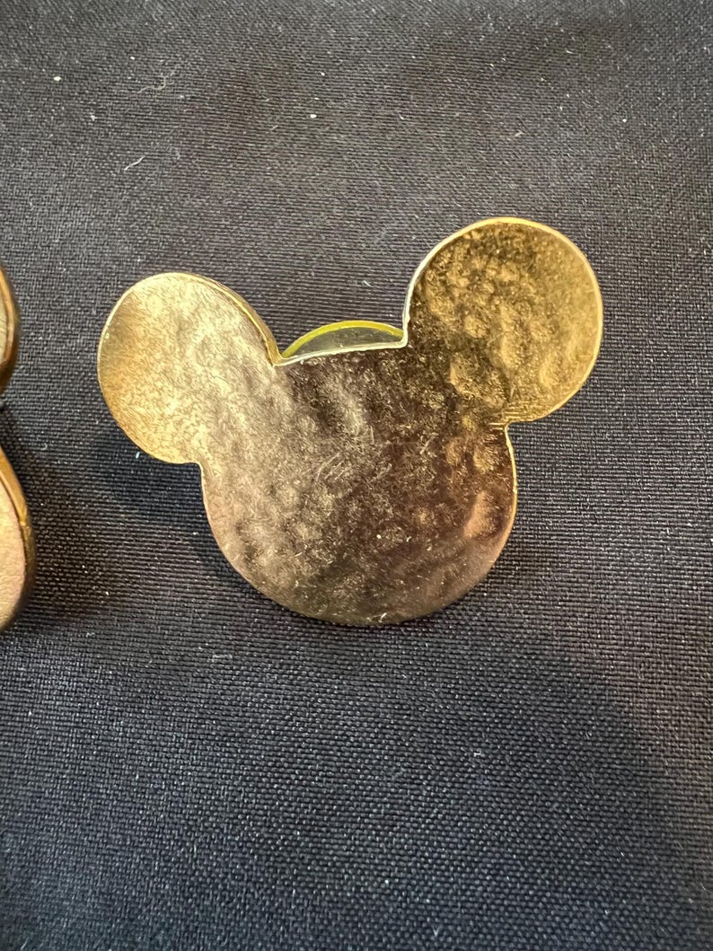 Pu&ograve; includere: Spilla dorata a forma di testa di Topolino con superficie strutturata. La spilla ha due orecchie circolari e una testa arrotondata. La spilla &egrave; su uno sfondo scuro.