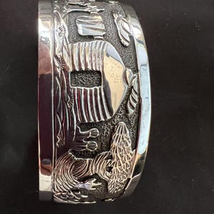 May include: A wide, silver-toned cuff bracelet with detailed relief carvings. The design includes stylized figures, possibly Native American-inspired, and natural elements. The bracelet has a polished finish and a curved shape, suitable for the wrist.
