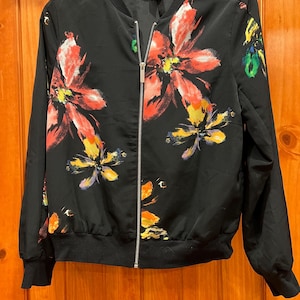 May include: Black bomber jacket with a floral print featuring pink, yellow, and green flowers. The jacket has a zipper closure and long sleeves.
