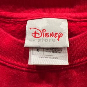 May include: A white tag with the text "Disney store" in red lettering. The tag also has the text "80% Cotton 20% Polyester Made in Mexico" and "Care on Reverse". The tag is attached to a red sweatshirt.