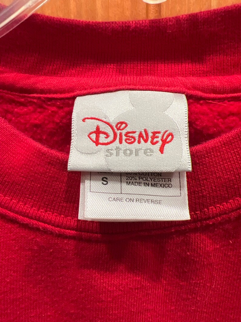 May include: A white tag with the text "Disney store" in red lettering. The tag also has the text "80% Cotton 20% Polyester Made in Mexico" and "Care on Reverse". The tag is attached to a red sweatshirt.